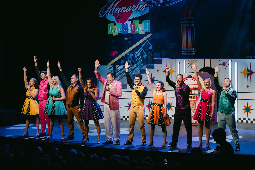 cast in colorful costumes with hand up and memories bandstand sign in background